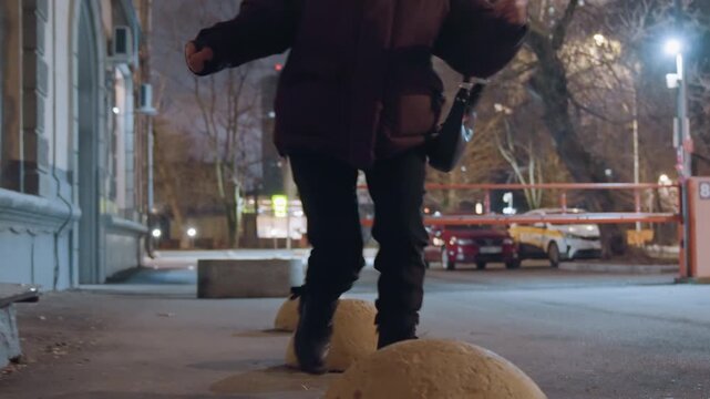Night street walker balancing on bollards, evening city lights, person in dark coat carrying bag stepping between yellow dome bollards, closeup boots tapping pavement, empty bench and parked car