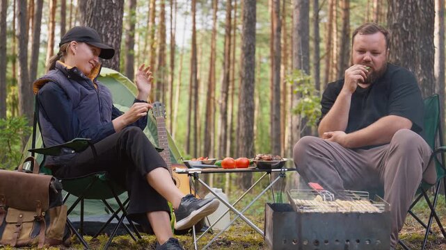 Young man and woman sitting in folding chairs, eating and talking while enjoying a relaxing picnic at their campsite with a tent and a smoking barbecue grill in a sunny pine forest