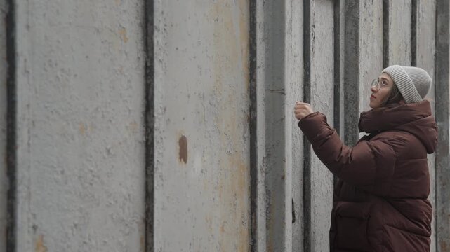 Person inspects tall concrete wall seam, wearing puffer jacket and knit hat while walking along rusted panel line urban street backdrop with parked cars, closeup on hands touching panel gap, mood