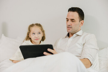 Fototapeta premium A father and daughter sit on a bed together, looking at a tablet, sharing a quiet moment indoors.