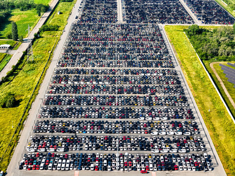 Aerial view of thousands of new cars parked in organized rows at a massive logistics hub surrounded by green fields and solar panels in Rivalta di Torino, Piedmont, Italy.