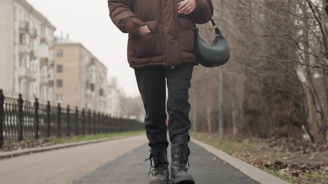 Closeup boots walking on asphalt path solitary commuter wearing brown puffer coat carrying shoulder bag, residential street with fence and bare trees, muted winter light, steady deliberate stride