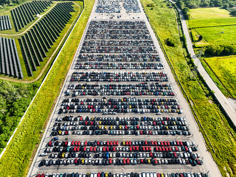 Aerial view of a massive car storage lot filled with rows of vehicles next to a large solar panel farm and green fields in Rivalta di Torino, Piedmont, Italy.