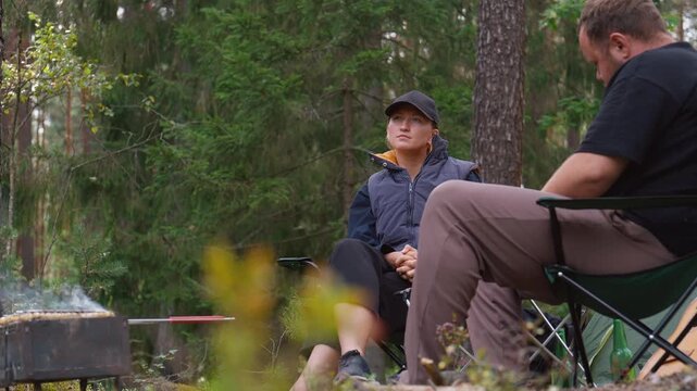 Young man and woman relaxing and talking at their campsite in a pine forest, drinking beer and grilling food on a barbecue as they enjoy their outdoor vacation together