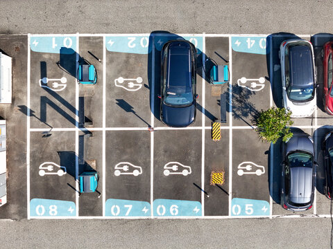 Aerial view of electric vehicle charging stations in a parking lot with numbered spots and parked cars under bright sunlight Turin, Piedmont, Italy.