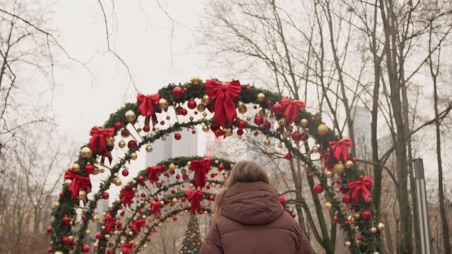 Girl with cheerful expressions strolling beneath colorful archways amidst urban scenery. Youthful woman engaging in playful movements while walking through seasonal tunnel with cityscape in background
