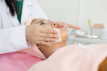Naklejka premium Young woman relaxes during facial treatment. Professional hands gently cleanse her face. Soft towel covers her head and shoulders. She closes eyes, appearing serene and calm 