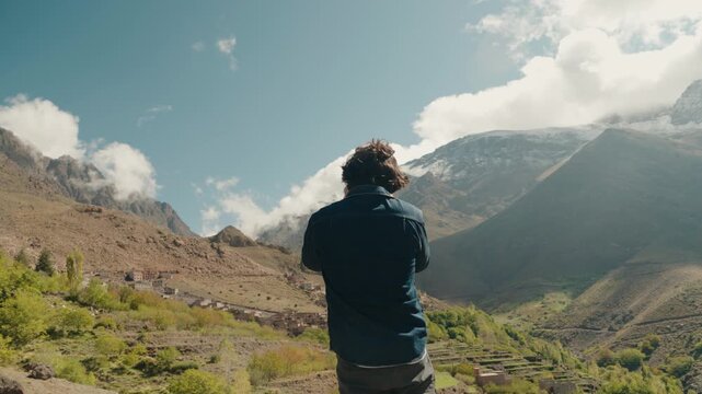 A man stands overlooking a beautiful Moroccan village nestled in the Atlas Mountains. The tranquil scene evokes a sense of wanderlust and adventure. He contemplates the stunning landscape.
