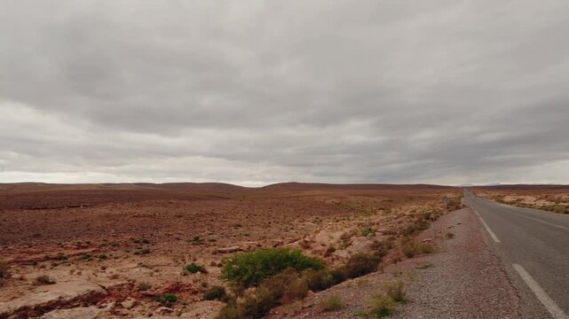 Vast desert landscape in Morocco under a cloudy sky. Reddish-brown terrain stretches to the horizon, sparse vegetation dots the area, and a quiet highway offers a sense of solitude. Perfect for travel