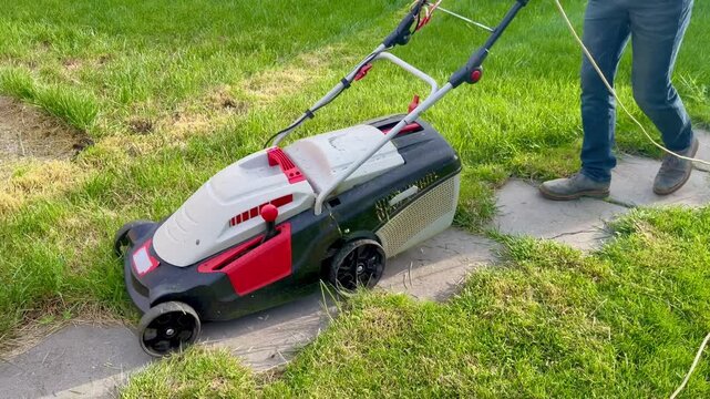 Man mowing lush green lawn with electric lawn mower on stone path in backyard garden on a sunny day
