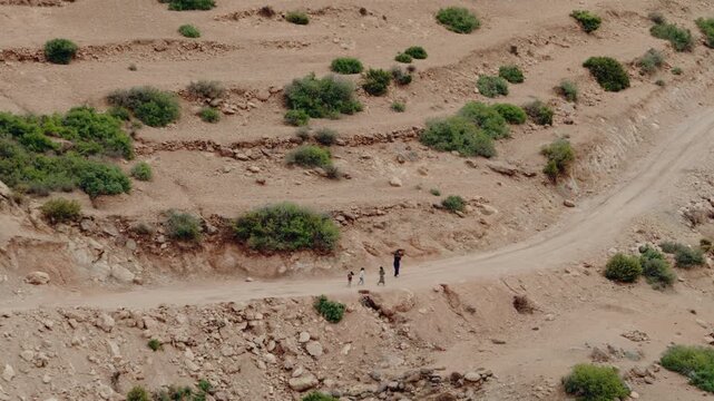 A woman and two children walk along a dusty road through a dry, rocky landscape in Morocco. They wear traditional clothing, and the woman carries a bundle on her head.