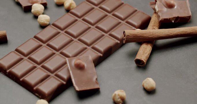 High-quality image of a chocolate bar surrounded by hazelnuts and cinnamon sticks on a dark surface. Perfect for food and dessert themes. Delicious Chocolate Bar with Hazelnuts and Cinnamon