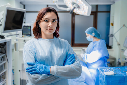 Portrait of female doctor looking at camera in operating room, blurred background with surgeons performing operation under surgical lights, professional healthcare surgery