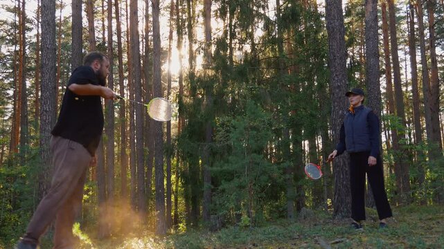A couple man and a woman are enjoying a game of badminton together in a beautiful pine forest, where golden sunlight breaks through the trees and accentuates their movements