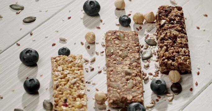 A variety of granola bars arranged on a white wooden table with scattered blueberries and nuts, showcasing a healthy snack option. Assorted Granola Bars with Blueberries and Nuts on White Wooden Table