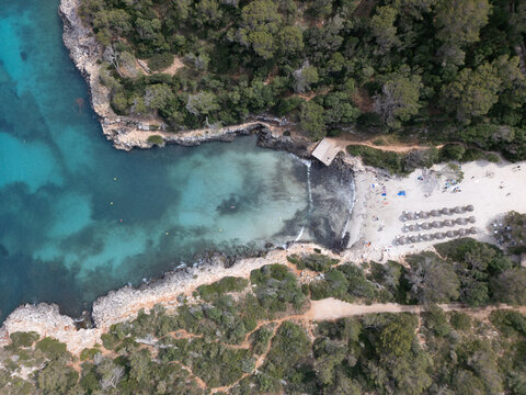 Aerial view of Cala Sa Nau turquoise waters, rocky cliffs, and a sandy beach with sunbeds surrounded by lush pine forests in Felanitx, Balearic Islands, Spain.