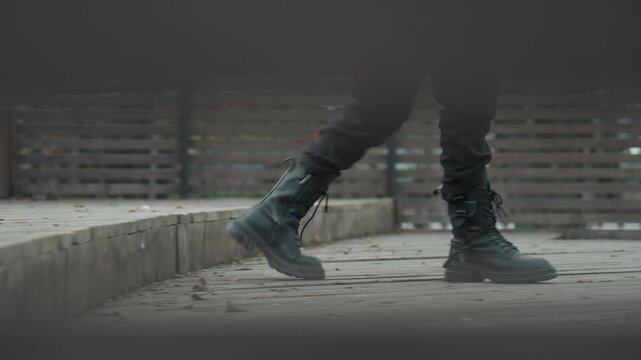 Closeup boots walking on boardwalk, gritty overcast mood, leather scuff on wooden planks, scattered leaves, lowangle perspective, slow deliberate pace, subtle motion blur, cinematic tone