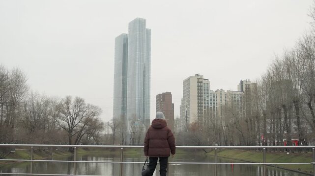 Woman at riverside railing overlooking pond, bundled in maroon coat, hands resting on metal rail, urban skyline reflected in still water, bare winter trees framing scene, local taking quiet pause