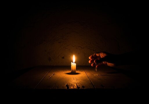 A single lit candle burning on a dark wooden table with a human hand reaching towards the flame in a moody atmosphere
