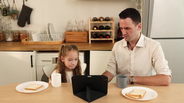 A father and daughter sitting at a kitchen table, engaging with a tablet device during breakfast with slices of bread and mugs in front of them.