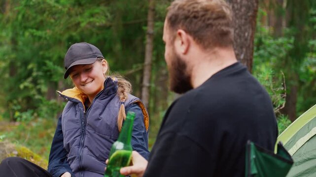 Young couple enjoying a picnic in the woods, with the man drinking beer from a green bottle and the woman eating and laughing while sitting together on a camping trip in a beautiful green forest