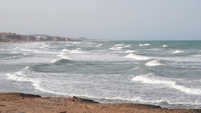 Strong sea waves rolling along Mediterranean coastline with urban beachscape and cloudy sky in Spain during windy day.