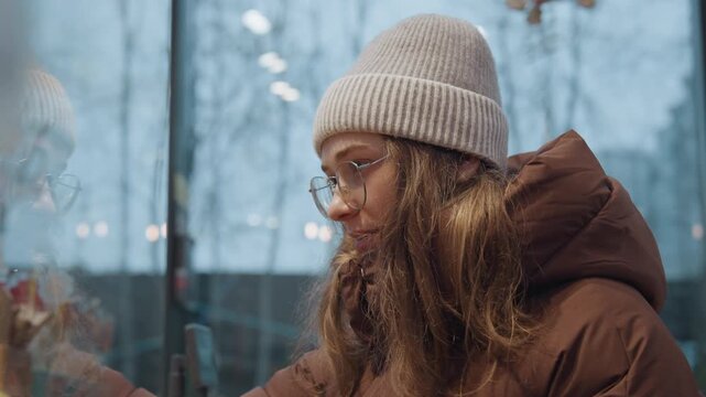 Reflected white woman in holiday window touching glass and studying gift display, wearing glasses and beanie, layered reflections and city lights create intimate atmosphere frames show gestures