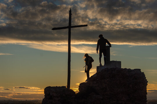 Silhouettes of couple beside cross on rock at sunset