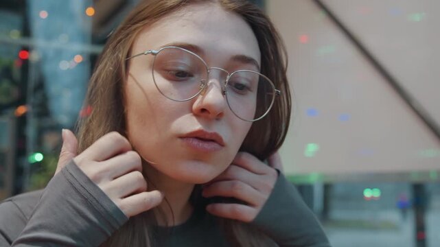 White woman adjusting glasses, pensive indoor portrait under colored lights, fingers touching hair and forehead, intimate closeup with reflective window, urban commuter moment and quiet introspection