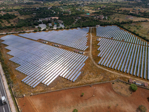 Aerial view of a large solar panel farm with rows of photovoltaic modules surrounded by rural fields and green trees in Manacor, Balearic Islands, Spain.