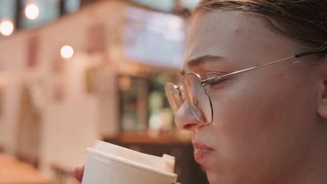 Closeup of woman sipping warm coffee in inviting cafe. Portrait of lady tasting latte amidst cozy cafe ambiance. Elegant woman with round glasses relishing warm beverage in charming cafe atmosphere