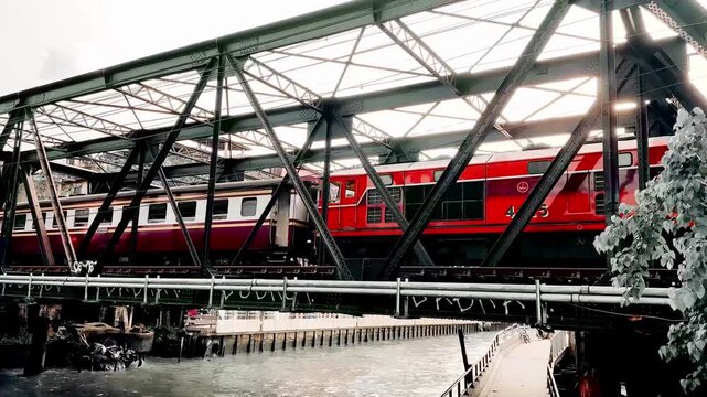 A red train crosses a steel railway bridge above an urban canal, capturing industrial architecture, city transportation, and everyday movement in a busy metropolitan environment.