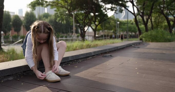 Portrait of junior schoolgirl with pigtails fastens velcro on shoe in park. Little girl primary school student adjusts sneaker as sits on stone parapet