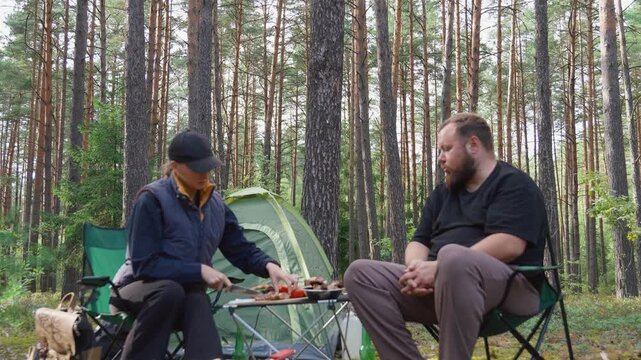 Young couple enjoying a picnic while sitting on chairs in a beautiful pine forest, with the woman slicing bread and the man taking vegetables from the table next to their tent and barbecue
