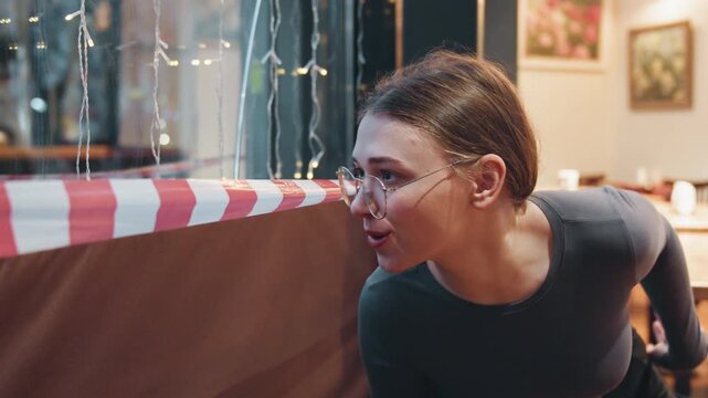 Caucasian woman conversing across caution tape, animated gestures and warm smile, seated at empty cafe booth, cozy lights and interior decor, candid reaction and questioning posture.