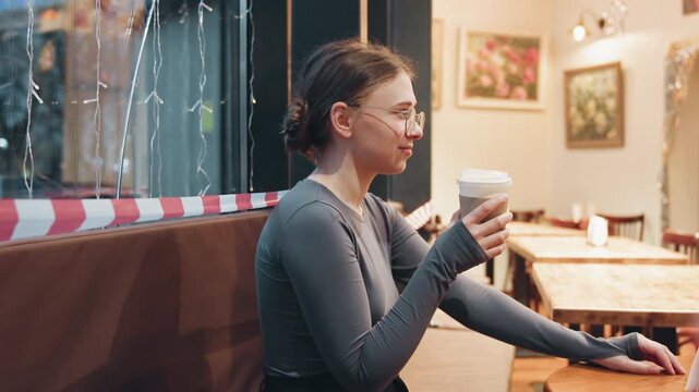Caucasian woman sipping coffee in empty cafe, seated in wooden booth with takeaway cup, contemplative gaze toward window, warm ambient lights and soft bokeh, quiet moment of solitude and nostalgia.