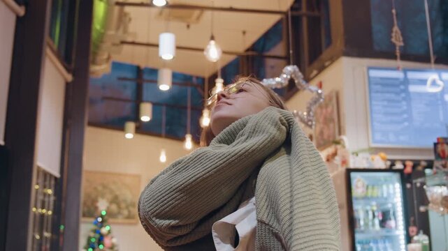 Young caucasian woman in cafe browsing menu and trying on scarf, glasses reflecting warm lights, counter and barista in background, sweater layers, thoughtful expression, seasonal holiday decor,