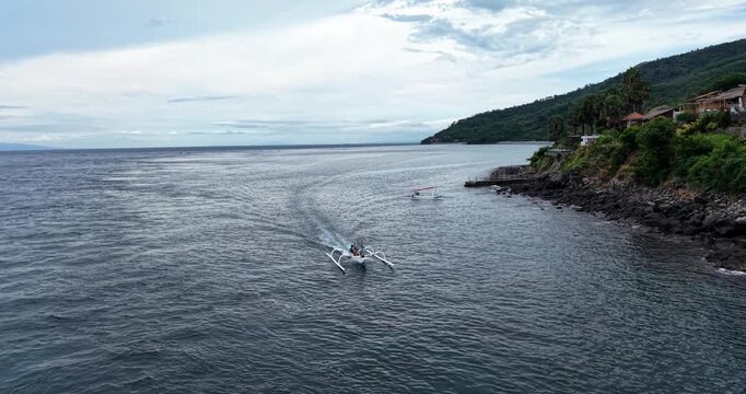 Drone aerial view of a traditional Balinese jukung boat cruising along the rocky tropical coastline of Amed, Bali, Indonesia with calm ocean waters and lush green hills.