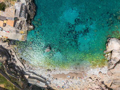 Aerial view of Cala Deia with clear turquoise water, rugged rocky cliffs, and people relaxing on the pebble beach Spain.