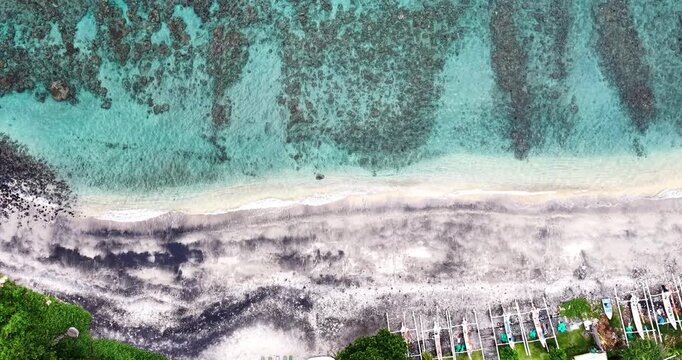 Top-down aerial drone footage of the volcanic black sand beach and turquoise coral reef waters in Amed, Bali, Indonesia with traditional fishing boats lined along the shoreline.