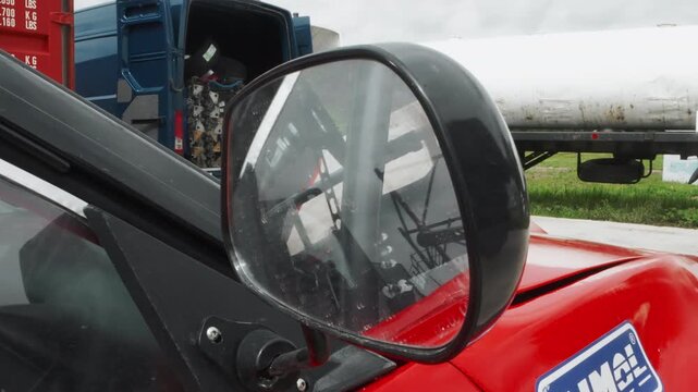 Closeup red vehicle mirror showing yard reflection and truck cab, roadside logistic backdrop, technician performing safety inspection, rain droplets on glass, chrome mount and bolts visible,