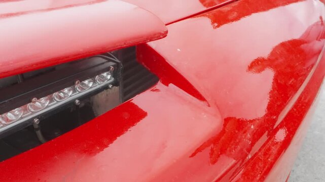 Closeup red car headlight showcasing led cluster, glossy paint reflections, rainspeckled surface, aerodynamic bumper and hood contours, photographer silhouette on body, tuning workshop atmosphere,