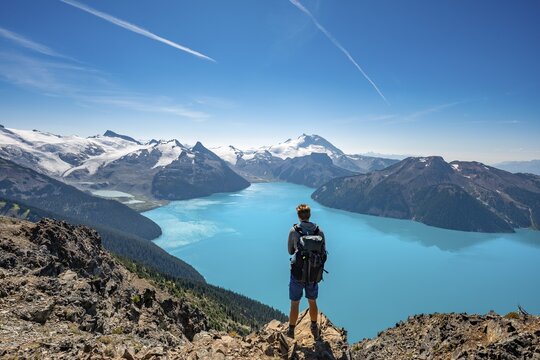 Young man standing on a rock, looking into the distance, view of mountains and glacier with turquoise blue lake Garibaldi Lake, peaks Panorama Ridge, Guard Mountain and Deception Peak, Garibaldi Provincial Park, British Columbia, Canada