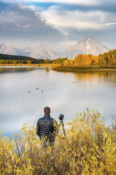 Photographer standing with tripod at the river, Snake River, Mount Moran at the back, morning mood at Oxbow Bend, autumn trees, Grand Teton Range, Grand Teton National Park, Wyoming, USA