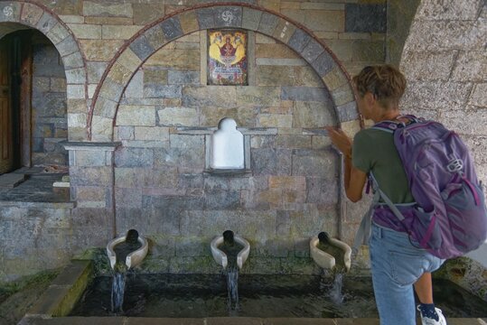A woman stands in front of the old stone fountain with three water sources and marble basin below the Panag&iacute;a church and looks at a mural, Mesochori, Karpathos, South Aegean, Greece