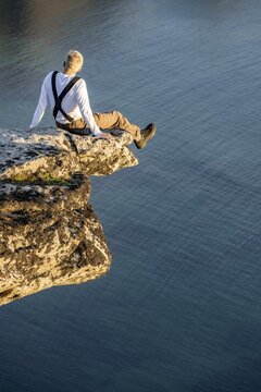 Hiker sitting on rock ledge on precipice above sea, summit of M&aring;tinden mountain, near Stave, Nordland, Norway