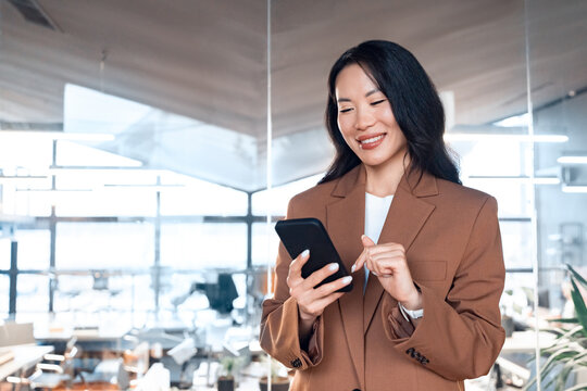 Middle-aged Asian business manager ceo woman using cell phone mobile app. Smiling mature businesswoman entrepreneur holding smartphone standing in office workplace working online on gadget. Copy space