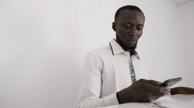Black man checking phone at table, white shirt and patterned tie, scrolling romantic messages with anxious smile, soft interior light, minimalist countertop, quiet evening anticipation and hopeful