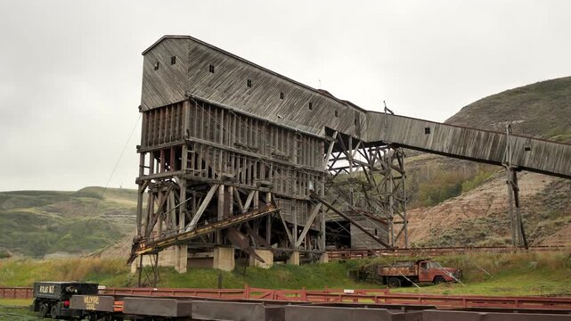 Wooden coal tipple at Drumheller, weathered timber structure with idle flatcars and rusting truck under overcast sky, long conveyor bridge stretching to hillside, grassy badlands Drumheller, Canada