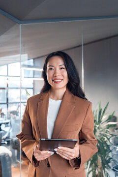 Vertical portrait middle-aged specialist business lady standing holding digital tablet at work in office, smiling at camera. Mature Asian professional manager woman leader entrepreneur using device